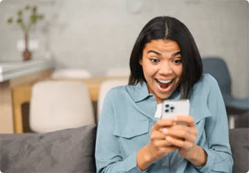  Eine Frau im blauen Hemd sitzt auf einem Sofa und hält ein Mobiltelefon. Sie hat ein weites Lächeln im Gesicht.