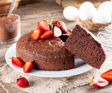 Ein Schokoladenkuchen aus dem ein Stück ausgeschnitten ist mit Erdbeeren auf einem Teller. Im Hintergrund sind frische Eier und Kakopulver.