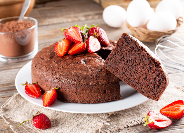 Ein Schokoladenkuchen aus dem ein Stück ausgeschnitten ist mit Erdbeeren auf einem Teller. Im Hintergrund sind frische Eier und Kakopulver.