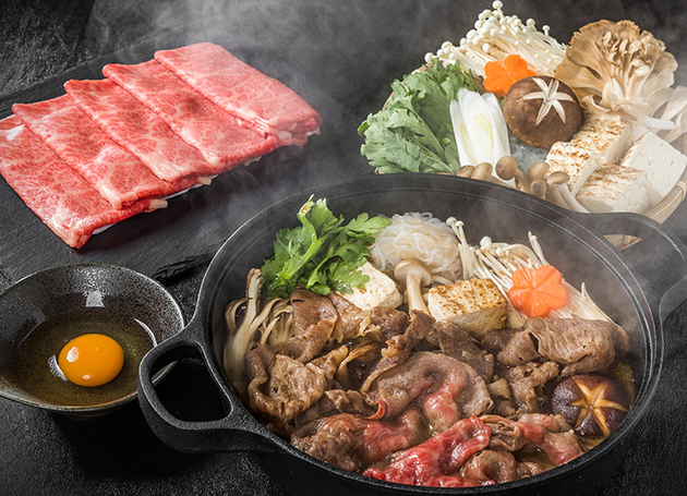 Eine Pfanne mit dampfendem hauchdünn geschnittenem Rindfleisch, Pilzen und Koriander. Im Hintergrund sind weitere frische Zutaten. 