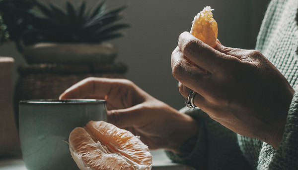 Eine Frau hält eine geschälte Orange in der Hand, vor ihr steht eine Tasse mit Tee.