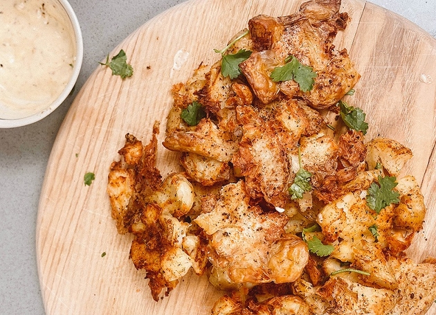 Gebratene zerschlagene Kartoffeln auf einem Holzschneidebrett. Daneben ist eine Schüssel mit Knoblauch-Dip