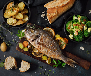 Ein gebratener Fisch und Gemüse auf einem Schneidebrett.Daneben sind Schüsseln mit Kartoffeln und Broccoli.