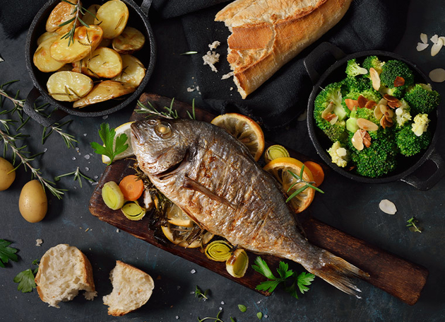 Ein gebratener Fisch und Gemüse auf einem Schneidebrett.Daneben sind Schüsseln mit Kartoffeln und Broccoli.