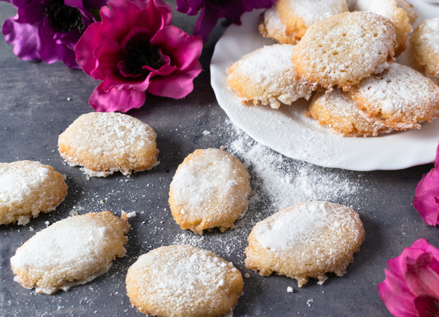 Mehrere Ricciarelli mir Puderzucker bestreut auf einem schwarzen Tisch, umgeben von rosa Blumen.