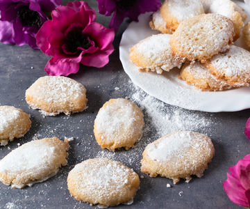 Mehrere Ricciarelli mir Puderzucker bestreut auf einem schwarzen Tisch, umgeben von rosa Blumen.