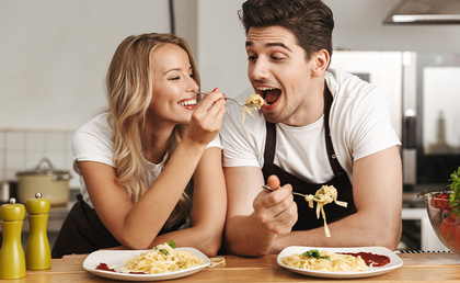 ein junger Mann und eine junge Frau mit Schürzen stehen am Küchentisch und essen Spaghetti von Tellern
