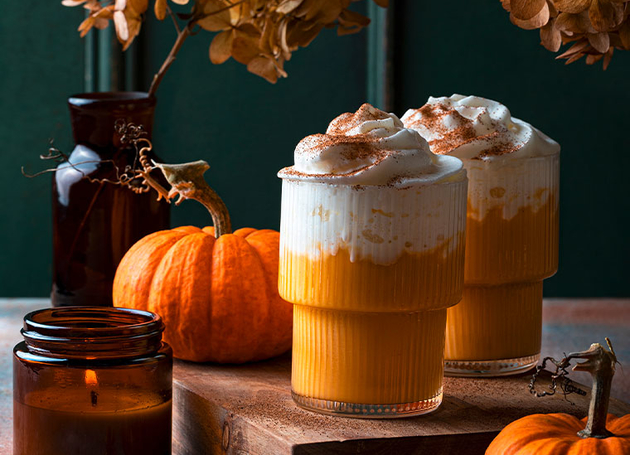 Zwei Glässer mir orangem Kürbiskaffee mit Schlagsahne obendrauf. Drumherum sind Herbstdekorationen wie Kürbise und eine Kerze.