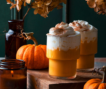 Zwei Glässer mir orangem Kürbiskaffee mit Schlagsahne obendrauf. Drumherum sind Herbstdekorationen wie Kürbise und eine Kerze.