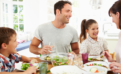 zwei Erwachsene und zwei Kinder sitzen lachend am Tisch und essen Gemüsesalat
