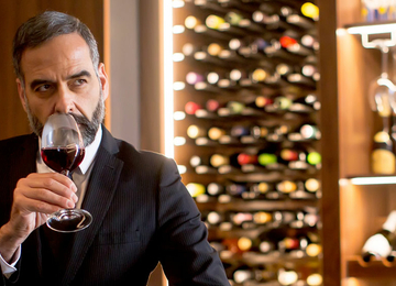 Ein gepflegeter Mann im Anzug richt an einem Glas Rotwein in einem Weinkeller.
