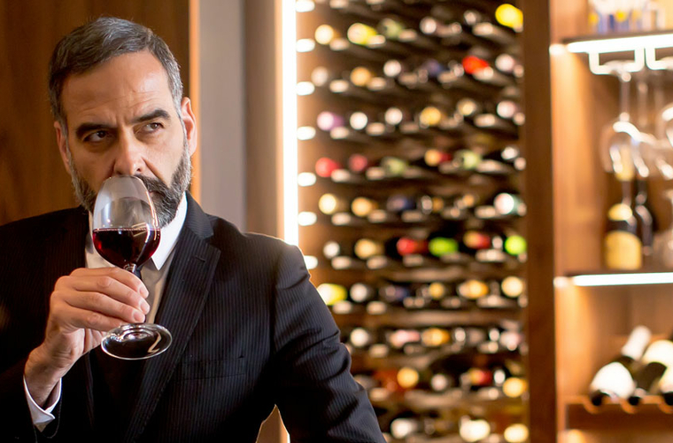 Ein gepflegeter Mann im Anzug richt an einem Glas Rotwein in einem Weinkeller.