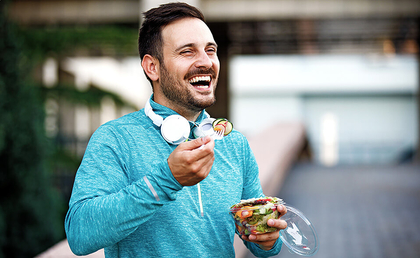ein junger lachender Mann mit Bart in einem blauen Sweatshirt isst einen kleinen Gemüsesalat aus einer Plastikschale