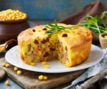 Ein Bohnen-Mais-Kuchen auf einem weissen Teller. Im Hintergrund ist eine Schüssel mit Maiskörnern.