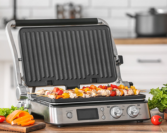 Ein offener Innengrill auf einer Küchentheke, gefüllt mit Spießen voller Gemüse und Fleisch. Um ihn herum sind gehackte Paprika, Salat, Brokkoli und Küchenutensilien zu sehen.