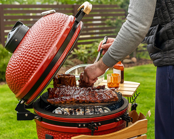Eine Person grillt Rippchen auf einem roten, runden BBQ-Smoker Grill im Freien. In der Nähe stehen eine Flasche Soße und Grillwerkzeuge.