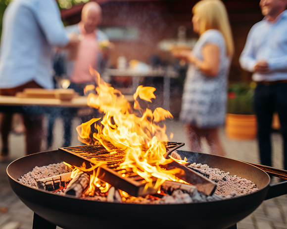 Ein brennender Außengrill mit sichtbaren Flammen im Vordergrund. Im Hintergrund unterhalten sich vier Personen und halten Getränke in der Hand, wobei eine Person ein gemustertes Kleid trägt.
