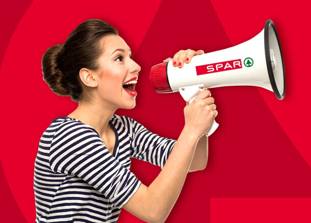 Dame in weiß-schwarzem gestreiftem T-Shirt mit kurzen Ärmeln hält ein Megafon mit SPAR-Logo und spricht hinein. Roter Hintergrund