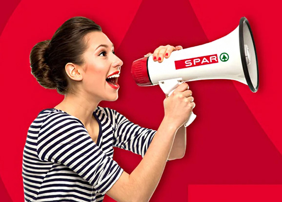 Dame in weiß-schwarzem gestreiftem T-Shirt mit kurzen Ärmeln hält ein Megafon mit SPAR-Logo und spricht hinein. Roter Hintergrund